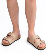 Beige double-strap slide sandals with cork-like midsoles and black outsoles, shown on a person's feet against a white background.