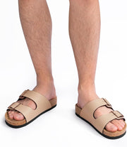 Beige double-strap slide sandals with cork-like midsoles and black outsoles, shown on a person's feet against a white background.