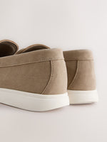 Close-up rear view of a pair of slip-on loafers in taupe suede with thick white soles, photographed on a clean light beige studio background.