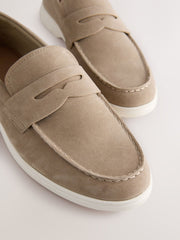 A pair of taupe suede penny loafers with white soles, shown in a close-up studio shot on a light beige background.