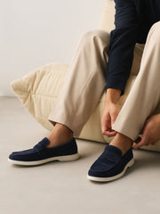 A person seated on a cream ribbed cushion wearing navy slip-on loafers with white soles and light beige trousers, adjusting one shoe in a clean indoor setting.