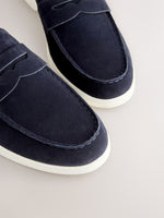 Close-up of a pair of navy blue suede penny loafers with white soles, shown on a light beige background.