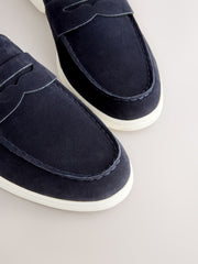 Close-up of a pair of navy blue suede penny loafers with white soles, shown on a light beige background.
